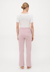 Vrouw met een bun, gekleed in een witte top met korte mouwen en lichtroze elastische broek, staan blootsvoets met de rug naar de camera op een witte achtergrond.