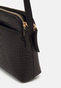 Czarna torebka z tłoczeniem imitującym skórę krokodyla, wyposażona w podwójne zamki błyskawiczne w złotym odcieniu oraz odpinany pasek. Miękka faktura, wyraźnie zarysowany kształt i szczegółowe przeszycia.