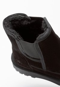 Čierne semišové členkové topánky s hustou fleecovou podšívkou a gumovou podrážkou, naklonené tak, aby ukázali interiér a bočný elastický panel pre dokonalé prispôsobenie.