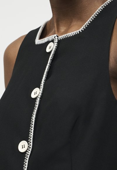 Μαύρο αμάνικο τοπ με λευκή ζιγκ-ζαγκ ραφή κατά μήκος των άκρων και τρία λευκά κουμπιά στην πρόσοψη, φορεμένο από ένα άτομο.