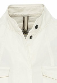 Biała kurtka z wysokim kołnierzem, ozdobiona ciemnymi guzikami oraz dwiema przednimi kieszeniami. Materiał wydaje się gładki z przeszytymi detalami.