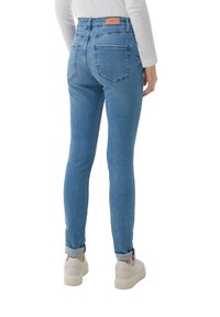 Jean skinny en denim bleu avec taille haute, poches arrière visibles, et un patch en cuir à la taille. Associé à des baskets de couleur claire.