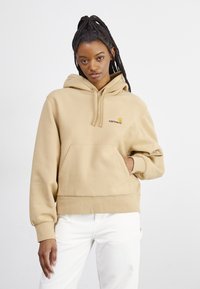 Beige hooded sweatshirt with a kangaroo pocket and ribbed cuffs. Features a small yellow and red logo on the chest. Soft fabric texture.