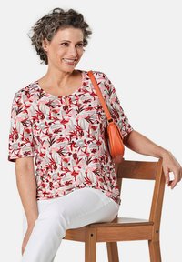 Bloes met korte mouwen in een bloemenpatroon in tinten roze, rood en grijs, voorzien van een ronde halslijn en een kleine keyhole-detail. Gecombineerd met witte broek.