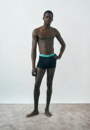 Άνδρας μοντέλο στέκεται σε δαπέδου με χαλί, φοράει μαύρα boxer briefs Nike με πράσινη ζώνη μέσης, ένα χέρι στη μέση, ουδέτερη έκφραση, απλό φωτεινό φόντο.