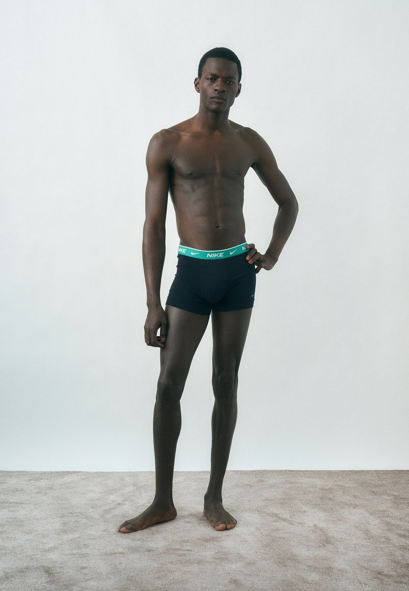 Άνδρας μοντέλο στέκεται σε δαπέδου με χαλί, φοράει μαύρα boxer briefs Nike με πράσινη ζώνη μέσης, ένα χέρι στη μέση, ουδέτερη έκφραση, απλό φωτεινό φόντο.