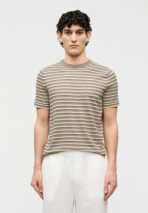 Νέος με κοντά κατσαρά μαλλιά φοράει κοντομάνικο πουκάμισο με μπεζ, μαύρες και λευκές ρίγες και λευκό παντελόνι, στέκεται μπροστά σε λευκό φόντο.