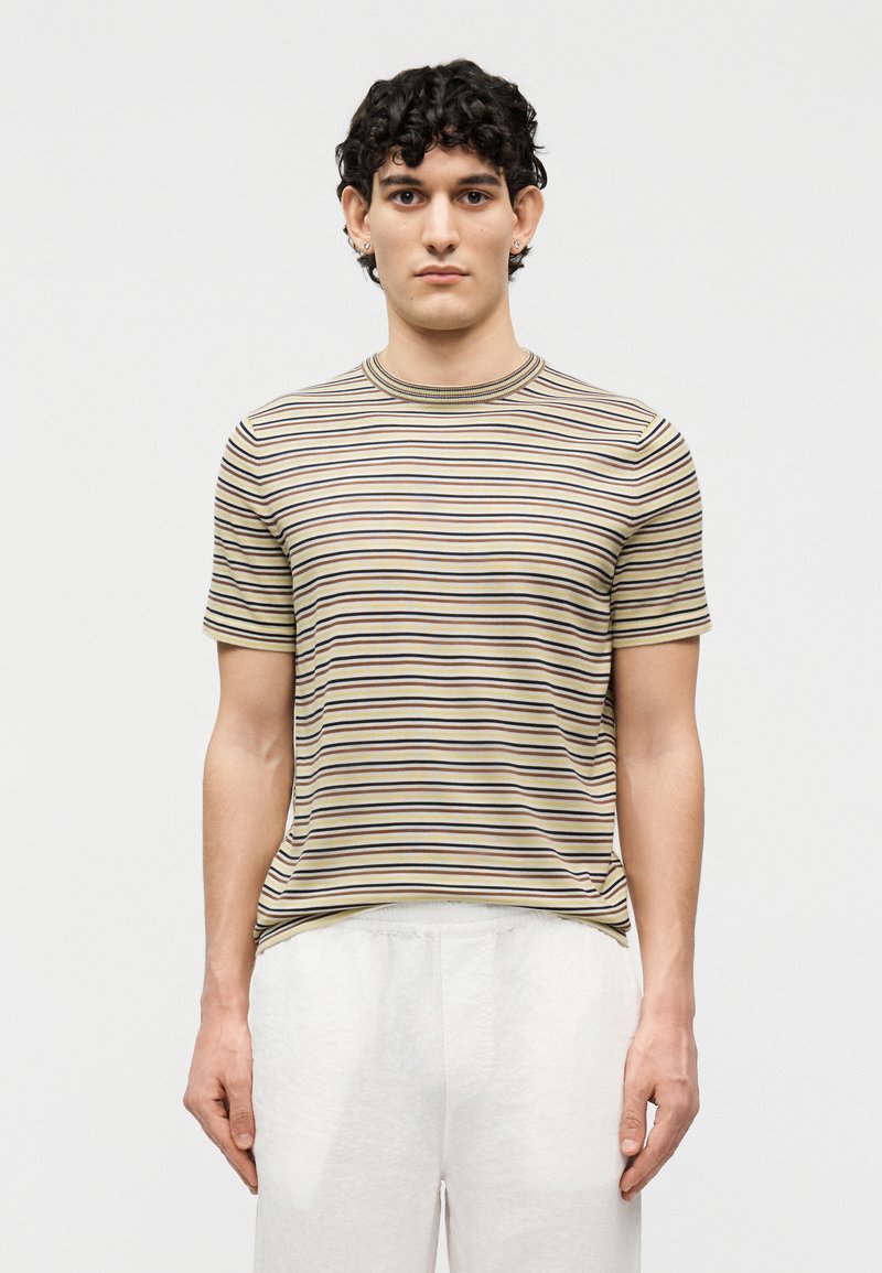 Νέος με κοντά κατσαρά μαλλιά φοράει κοντομάνικο πουκάμισο με μπεζ, μαύρες και λευκές ρίγες και λευκό παντελόνι, στέκεται μπροστά σε λευκό φόντο.