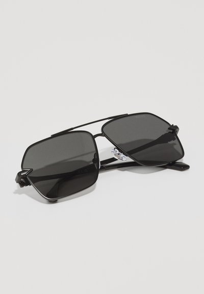 Lunettes de soleil noires avec une monture carrée, en métal, dotées de verres foncés et d'un design minimaliste. Les accents incluent des plaquettes de nez et des branches fines.