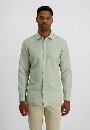 Man staat recht vooruit kijkend, draagt een lichtgroen overhemd met lange mouwen en beige broek tegen een effen achtergrond.