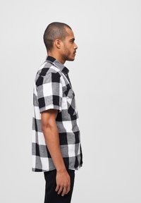 Korte mouwen, zwart-wit geblokt overhemd met een button-down kraag, voorzien van grijze accenten en twee voorzakken. Draagt over een zwarte broek.