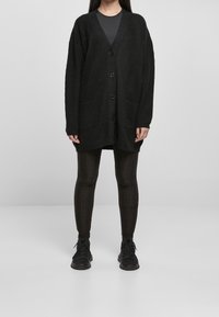 Cardigan noir en tricot avec une coupe surdimensionnée, poches avant et cinq boutons, présenté sur un modèle portant des leggings noirs texturés et des baskets.