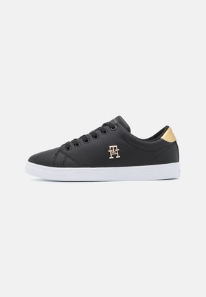 Zapatilla negra de corte bajo con suela blanca, cordones negros, lengüeta dorada en el talón y logo "TH" dorado en el lateral.