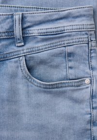 Zbliżenie na jasnoniebieskie jeansy z denimu, pokazujące przednią kieszeń, szlufkę na pasek, detale szwów oraz metalowy nit na fakturze tkaniny.