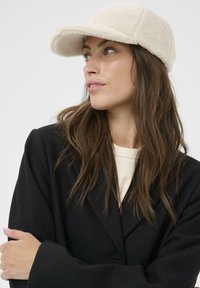 Faux fur cream cap with curved brim; cozy texture; six-panel design; paired with a black coat and light-colored shirt.