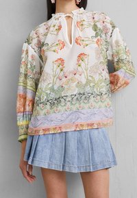 Blouse à imprimé floral blanche, avec un col en forme de trou, des manches longues et des accents colorés en rose, vert et lavande. Associée à une jupe en denim.
