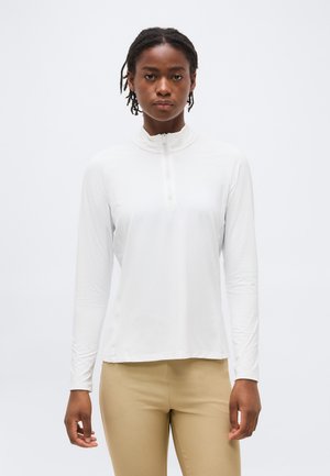 Žena s krátkymi dredmi, oblečená v bielom dlhorukávovom tričku so zipsom a béžových nohaviciach, stojaca na jednoduchom svetlom pozadí.