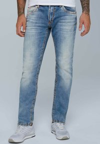 Jeans de mezclilla azul claro con corte ajustado, que presentan costuras en contraste, detalles desgastados y un ligero desvanecimiento. Combinados con zapatillas deportivas blancas.