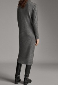 Szara dzianinowa sukienka z prążkowanej tkaniny, z długimi rękawami, sięgająca do kolan, z wysokim dekoltem i dopasowanym krojem. Modelka ma na sobie wysokie czarne kozaki.