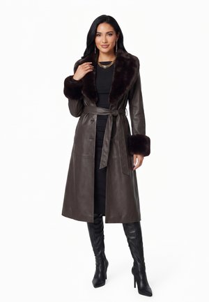 Donna che indossa un cappotto in pelle marrone scuro con colletto e polsini in pelliccia, vestito nero, stivali neri con tacco alto e gioielli d'oro, in piedi con la mano sul colletto.
