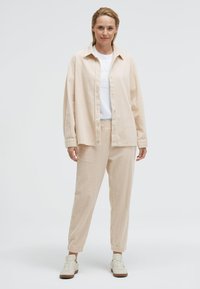 Beige strukturierte Hemd und passende Hose im entspannten Schnitt. Darunter ein weißes T-Shirt mit dezentem Logo, kombiniert mit hellen Sneakers.