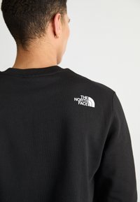 Sudadera negra con un cuello redondo y puños acanalados. Presenta un logo blanco de The North Face en la parte posterior, hecha de una tela suave y lisa.