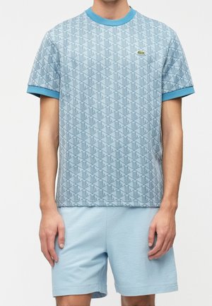 Mężczyzna w jasnoniebieskim T-shircie z geometrycznym wzorem oraz dopasowanych jednokolorowych jasnoniebieskich szortach, stojący na jednolitym tle.