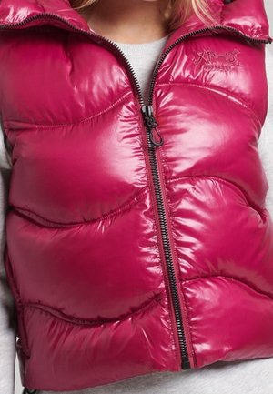 Close-up of a shiny magenta puffer vest with a central zipper worn over a light gray top, showing quilted stitching and embroidered logo.