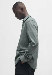 Lange mouwen met kraag in gemêleerd groene stof. Voorzien van een subtiel borstlogo en knoopsluiting aan de mouwen. Slim fit vorm, zachte textuur.