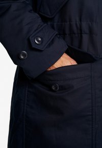 Granatowa kurtka o gładkim wykończeniu tkaniny. Zapinana na duże guziki na mankiecie i bocznej kieszeni, z wyraźnym trwałym szyciem i detalami wzorniczymi.