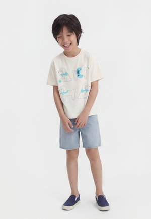 Garçon souriant portant un t-shirt crème avec quatre illustrations de caniches bleus, un short en denim clair et des chaussures bateau bleu marine, debout devant un fond blanc.