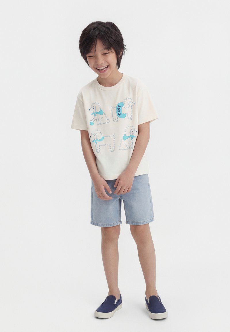 Χαμογελαστό αγόρι που φοράει κρεμ μπλουζάκι με τέσσερα μπλε σχέδια γαλλικού κόκερ, ανοιχτόχρωμα τζιν σορτς και μπλε παντοφλάκια, στέκεται μπροστά σε λευκό φόντο.