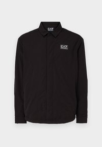 Svart jacka med krage, knappstängning, långa ärmar och en liten vit logo på bröstet. Slät och lätt tyg.