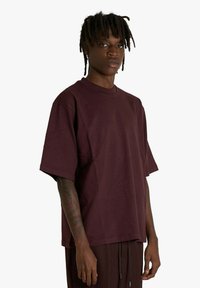 T-shirt in cotone bordeaux con maniche corte, collo rotondo e vestibilità rilassata, indossata da una modella, con un design semplice e senza marchi visibili.