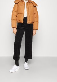 Doudoune marron avec un col haut et une fermeture éclair à l'avant, associée à un pantalon en velours côtelé noir cropped et des chaussures de sport blanches.