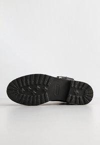 Semelle de bottes noire avec des crampons profonds et des motifs en forme de croix, vue de dessous sur un fond clair uni.