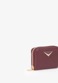 Portafoglio in pelle bordeaux con zip dorata, dettaglio hardware triangolare, forma quadrata e superficie liscia. Design compatto per carte e contanti.