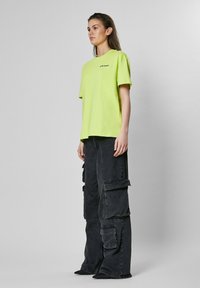 Lime zöld, oversized póló, pamut anyagból, rövid ujjú; fekete, bőszárú cargo nadrággal párosítva, nagy zsebekkel és megkopott textúrával.