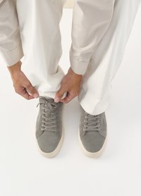 Γκρι σουέτ αθλητικά παπούτσια με ταιριαστά κορδόνια και λευκή καουτσούκ σόλα. Χέρια ατόμου ρυθμίζουν το παπούτσι κοντά σε ελεφαντόδοντο παντελόνι σε ανοιχτό φόντο.