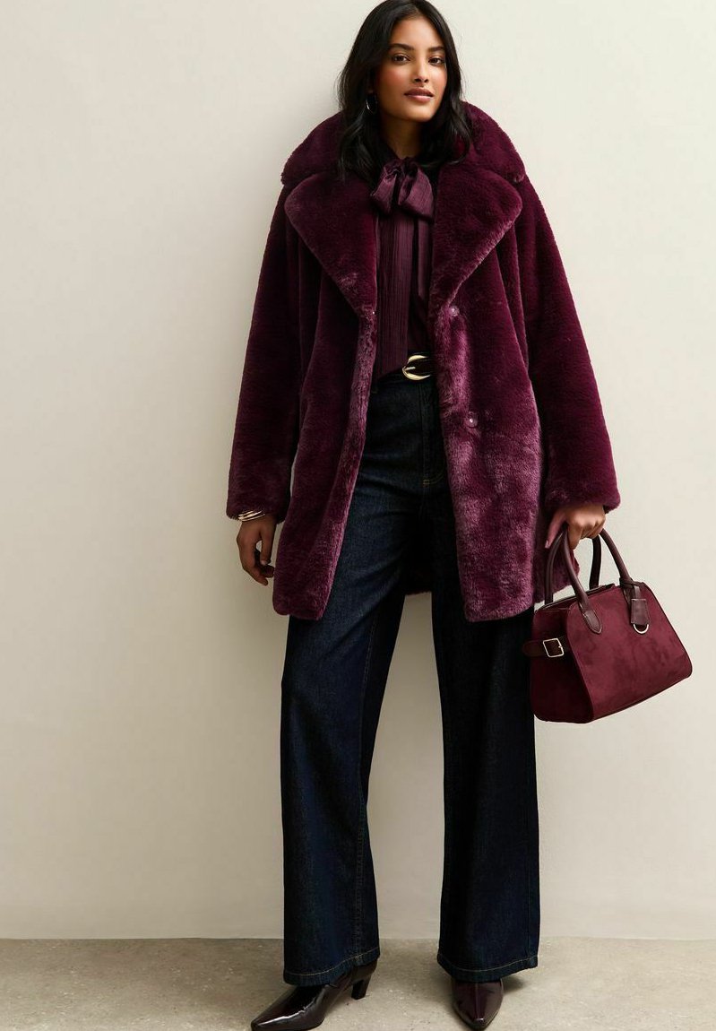 Mujer que viste un abrigo de pelaje sintético burdeos oscuro, blusa a juego, pantalones vaqueros de pierna ancha, botas de punta y sostiene un bolso burdeos.
