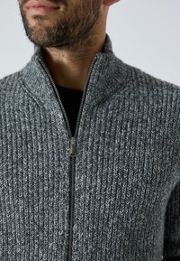 Grauer gestrickter Zip-Up-Pullover mit hohem Kragen, der eine gerippte Textur und einen silbernen Reißverschluss vorne aufweist.