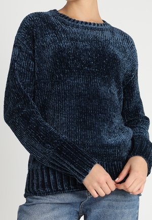 Personne portant un pull en velours côtelé bleu foncé à manches longues et un jean bleu, ajustant l'ourlet du pull.