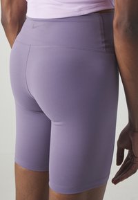 Personne portant un short de sport ajusté violet clair et un haut rose pâle, vue de dos de la taille jusqu'au milieu de la cuisse.