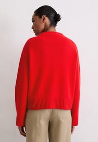 Czerwony sweter z dzianiny z ribbowym wykończeniem u dołu i na mankietach, o luźnym kroju i szerokim dekolcie, w połączeniu z beżowymi spodniami, pokazujący tylny widok.