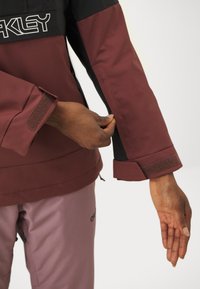 Veste bordeaux et noire au design élégant, dotée de poignets en Velcro et d'un matériau imperméable, présentée avec une manche partiellement déployée.