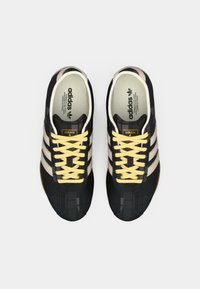 Black athletic shoes with yellow laces, featuring three horizontal tan stripes, textured surface, and a rounded toe design.