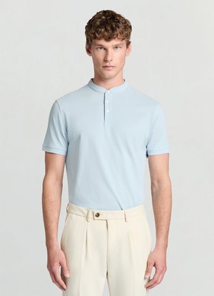 Polo azzurro chiaro a maniche corte con colletto a banda, caratterizzata da una placca con bottoni. Indossata con pantaloni sartoriali color crema.