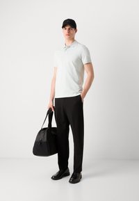 Světle zelené polo tričko, černé kalhoty a černé boty. Osoba drží černou sportovní tašku. Čepice má jemnou logo ozdobu.