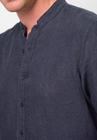 Camicia di lino blu navy con colletto rialzato, pattina frontale con bottoni, design minimalista e un tessuto morbido e strutturato.