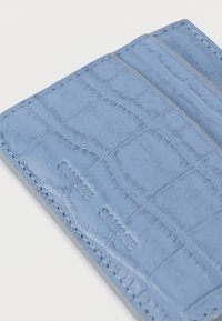 Modri usnjeni držalo za kartice z vzorcem krokodilje kože, opremljeno s številnimi režami za kartice in kontrastnim šivom. Logotip blagovne znamke vrezan na površino.